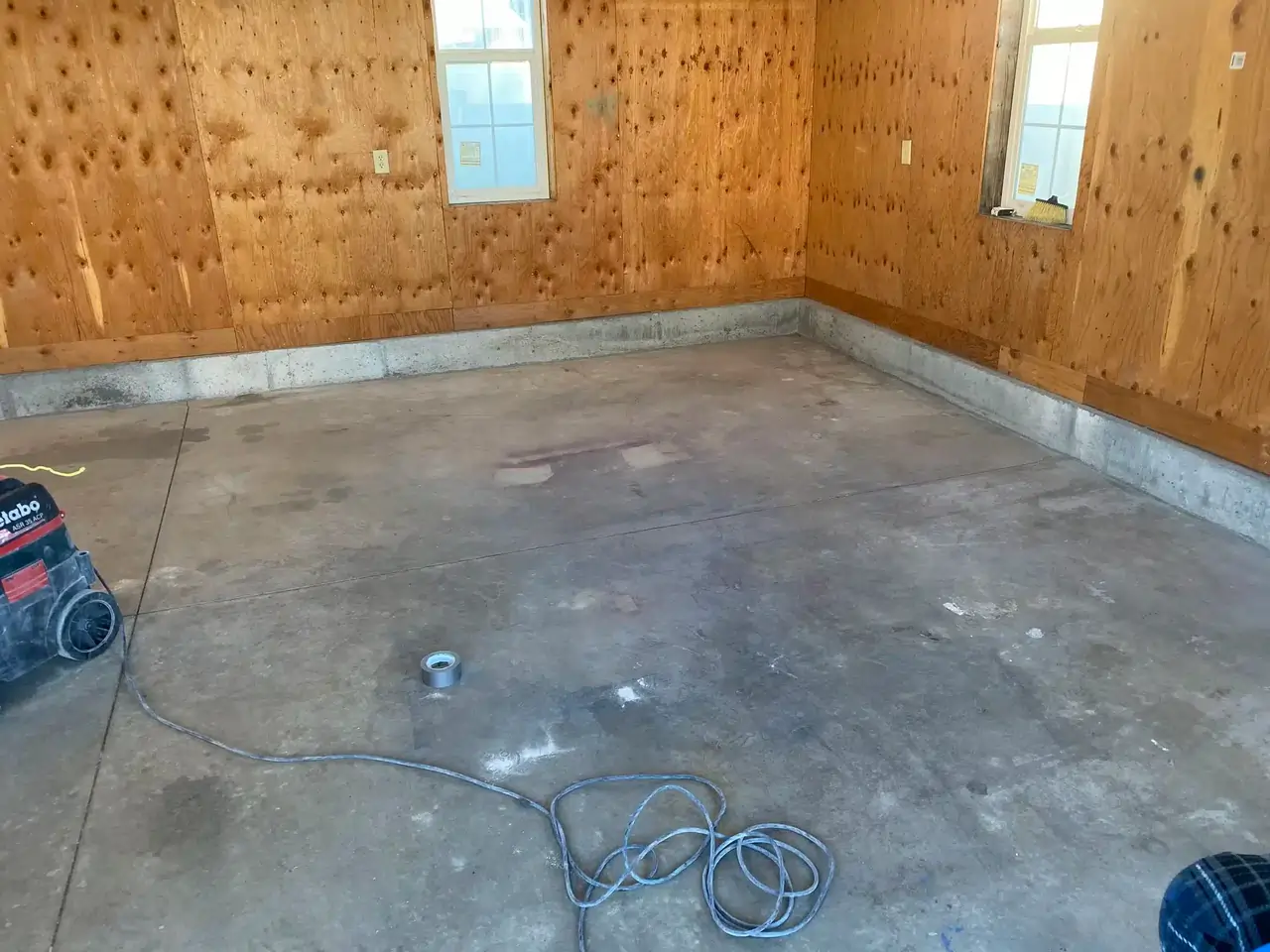 A before photo of a brown room with an unpolished concrete tiled floor and a wired equipment on the side.
