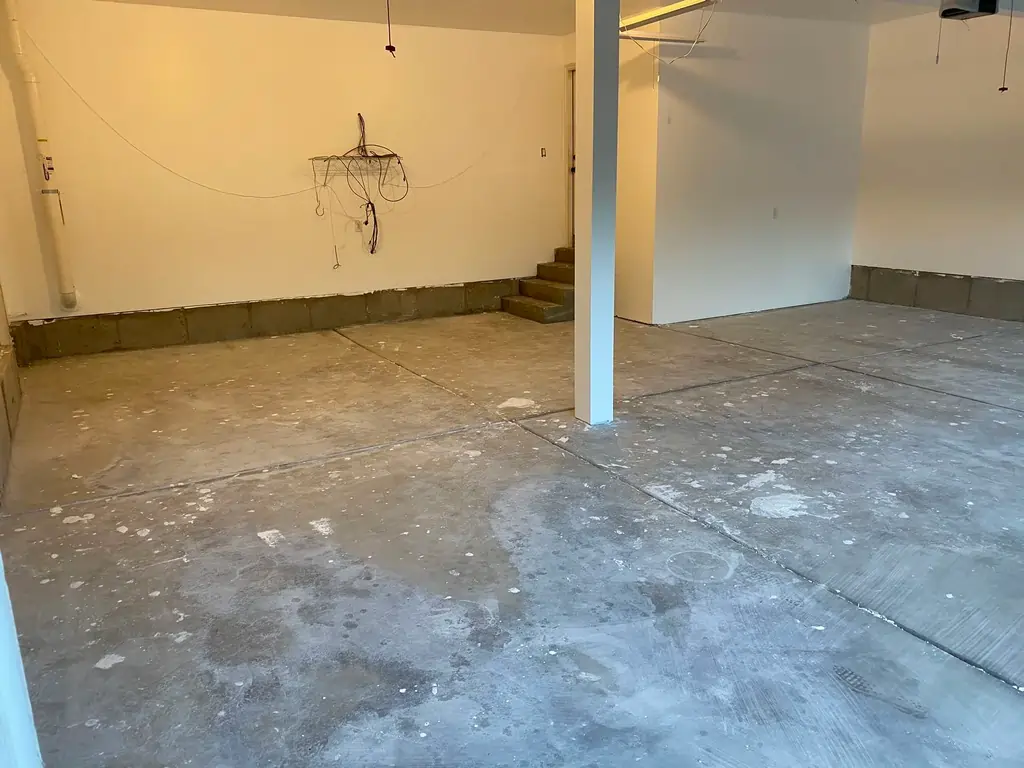 A before photo of a room that looks unfinished with a chipped concrete floor and exposed pipes on the wall