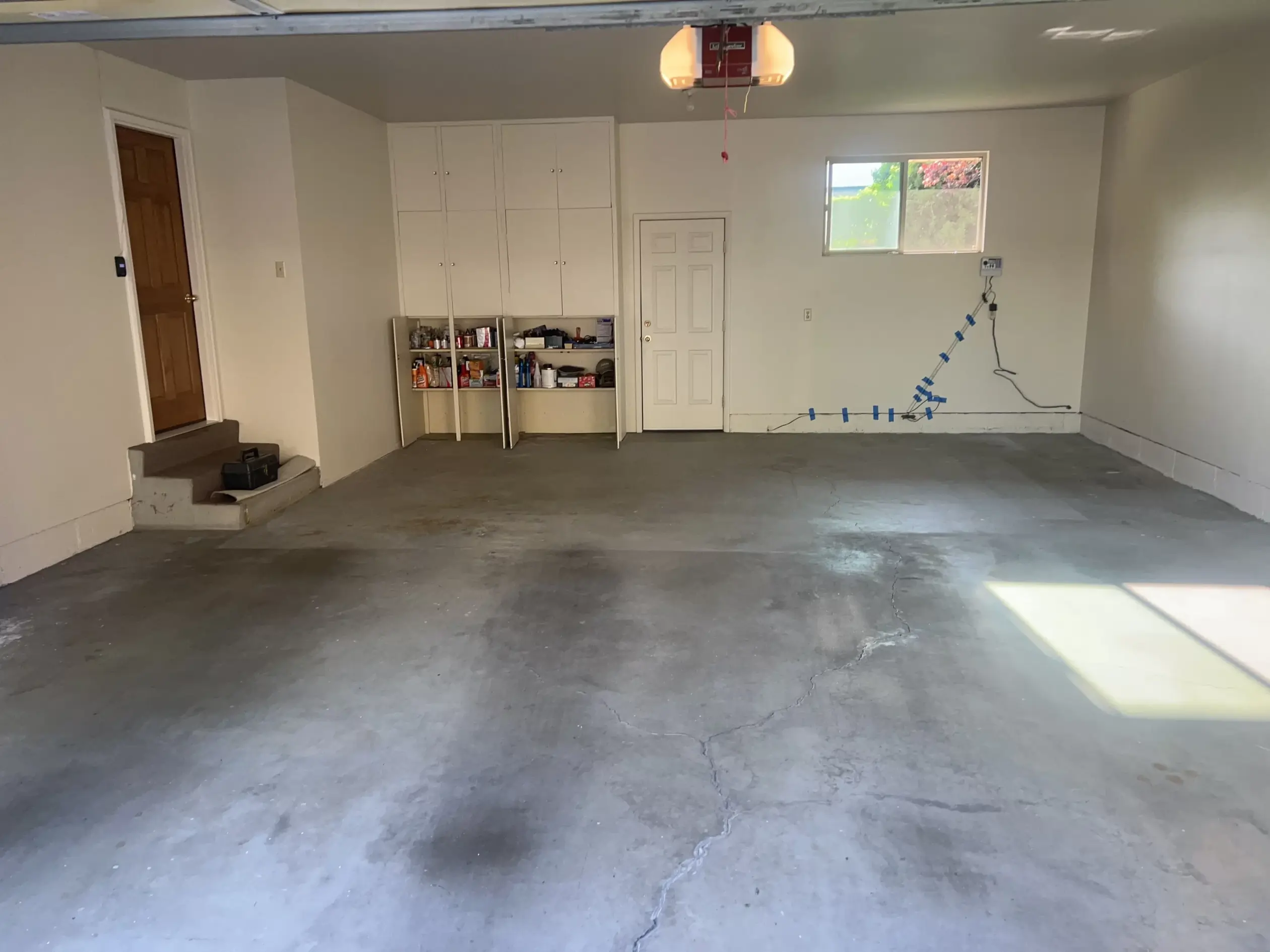 A before photo of an empty garage with an unpolished floor, white shelves, and brown door.